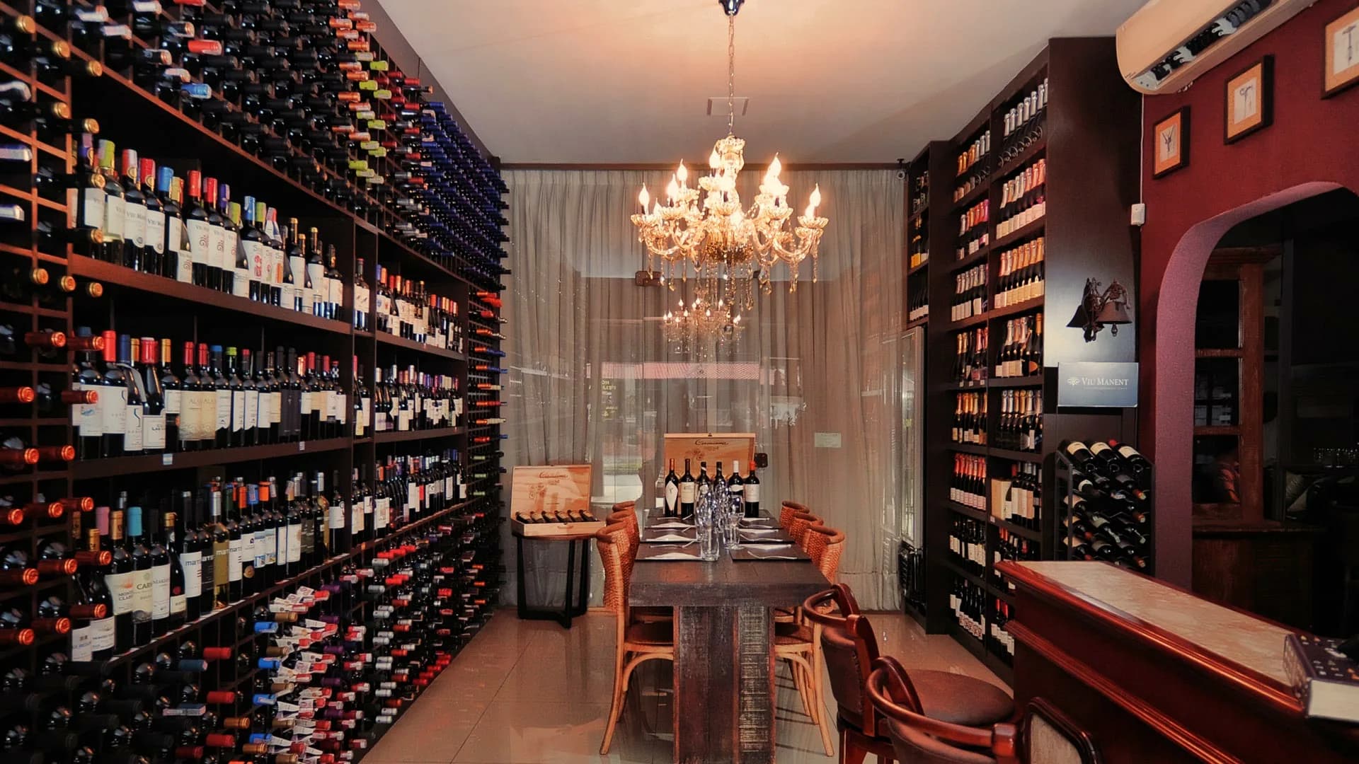 Adega do Empório Confrades com prateleiras de vinhos, lustre de cristal e mesa comunitária posta